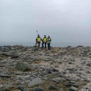 Chris Maio, Chris Tran, and Harper Baldwin stand next to each other, centered on a rocky shore horizon. Chris Tran is holding a Trimble GPS instrument
