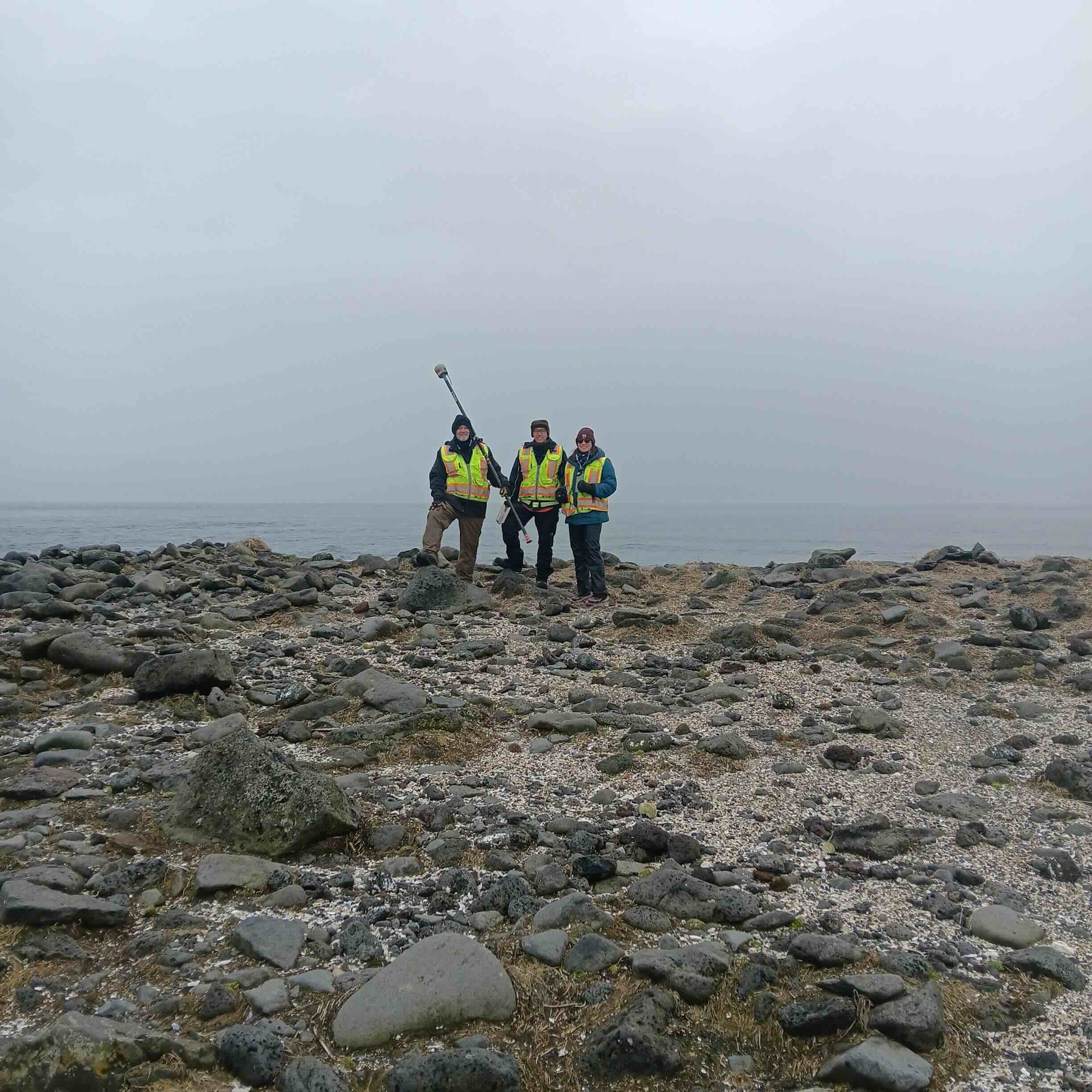 Chris Maio, Chris Tran, and Harper Baldwin stand next to each other, centered on a rocky shore horizon. Chris Tran is holding a Trimble GPS instrument