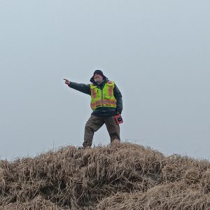 Chris stands silhouetted on top of a grassy hill against a grey overcast sky. He points at something out of frame.