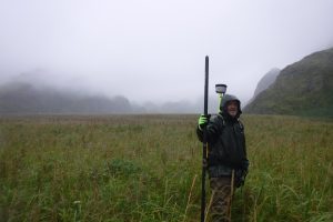 Chris is standing with a black pole in a grassy field, the background shows foothills and fog