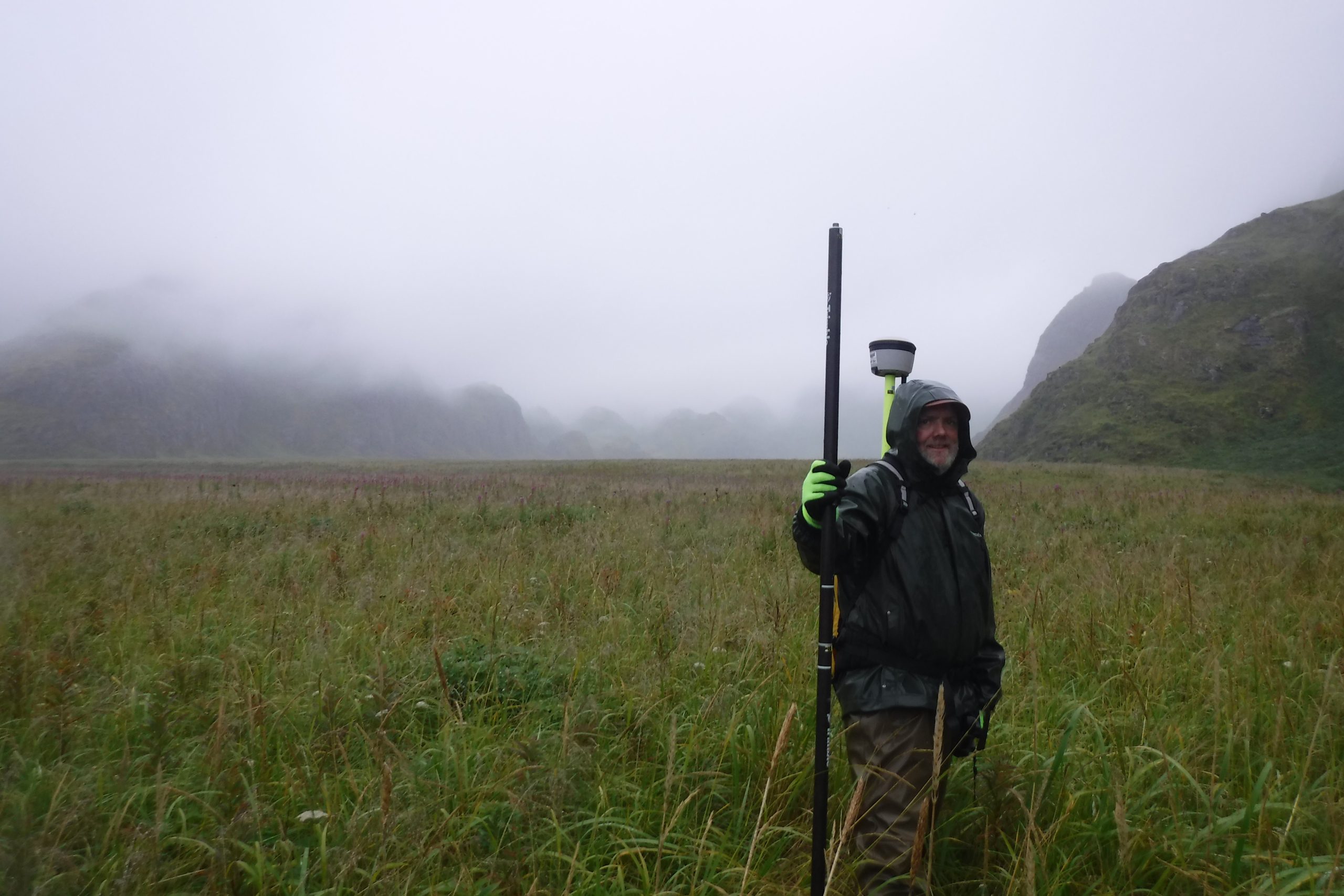 Chris is standing with a black pole in a grassy field, the background shows foothills and fog