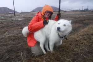 A white, fluffy, husky-like dog is snuggled up to someone and happily receiving but scratches. They are in a flat grassy field, there is a town further in the distance behind them and two hills. The sky is overcast.