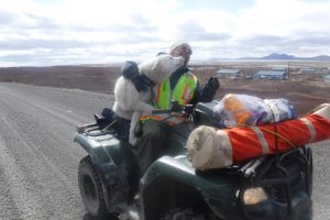 A fluffy white dog sits on an ATV with a person. The dog is not-so-subtly trying to steal a smooch from the person, the person is leaning away from the smooch. They are on a gravel road, some houses sit in a grassy field behind them and the sky is cloudy.