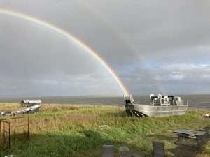 A metal boat sits on a grassy hill with a double rainbow positioned on top of the boat, the ocean is in the background