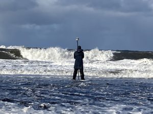 Someone stands in shallow water, facing away from the camera, and holding a Trimble GPS device. The sky is cloudy and there are large waves crashing in the background of the image.