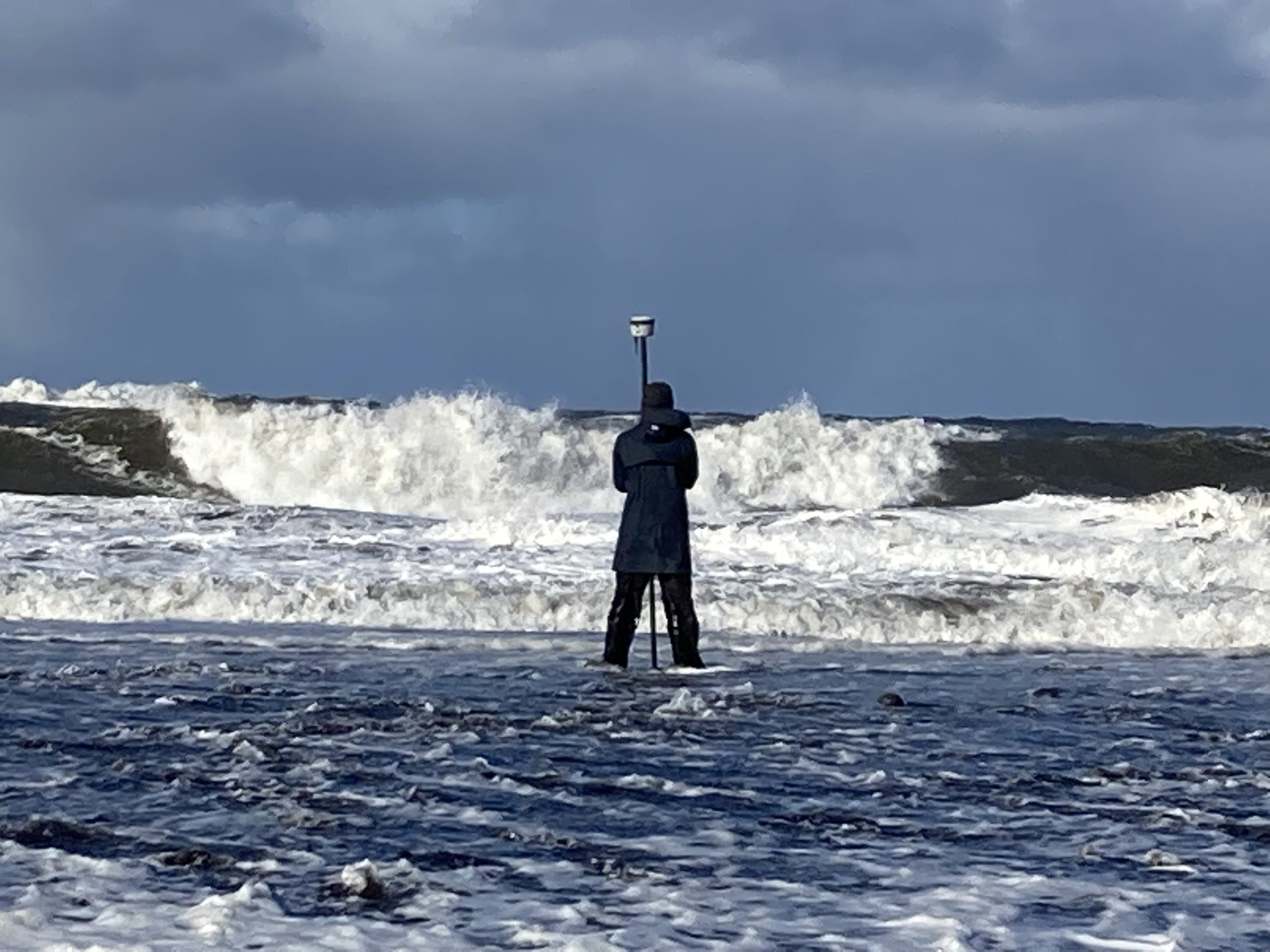 Someone stands in shallow water, facing away from the camera, and holding a Trimble GPS device. The sky is cloudy and there are large waves crashing in the background of the image.