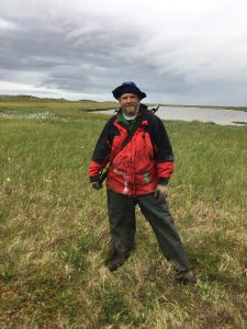 Chris stands in a grassy field with white flowers speckled througout the background, as well as a small pond in the background and a cloudy sky