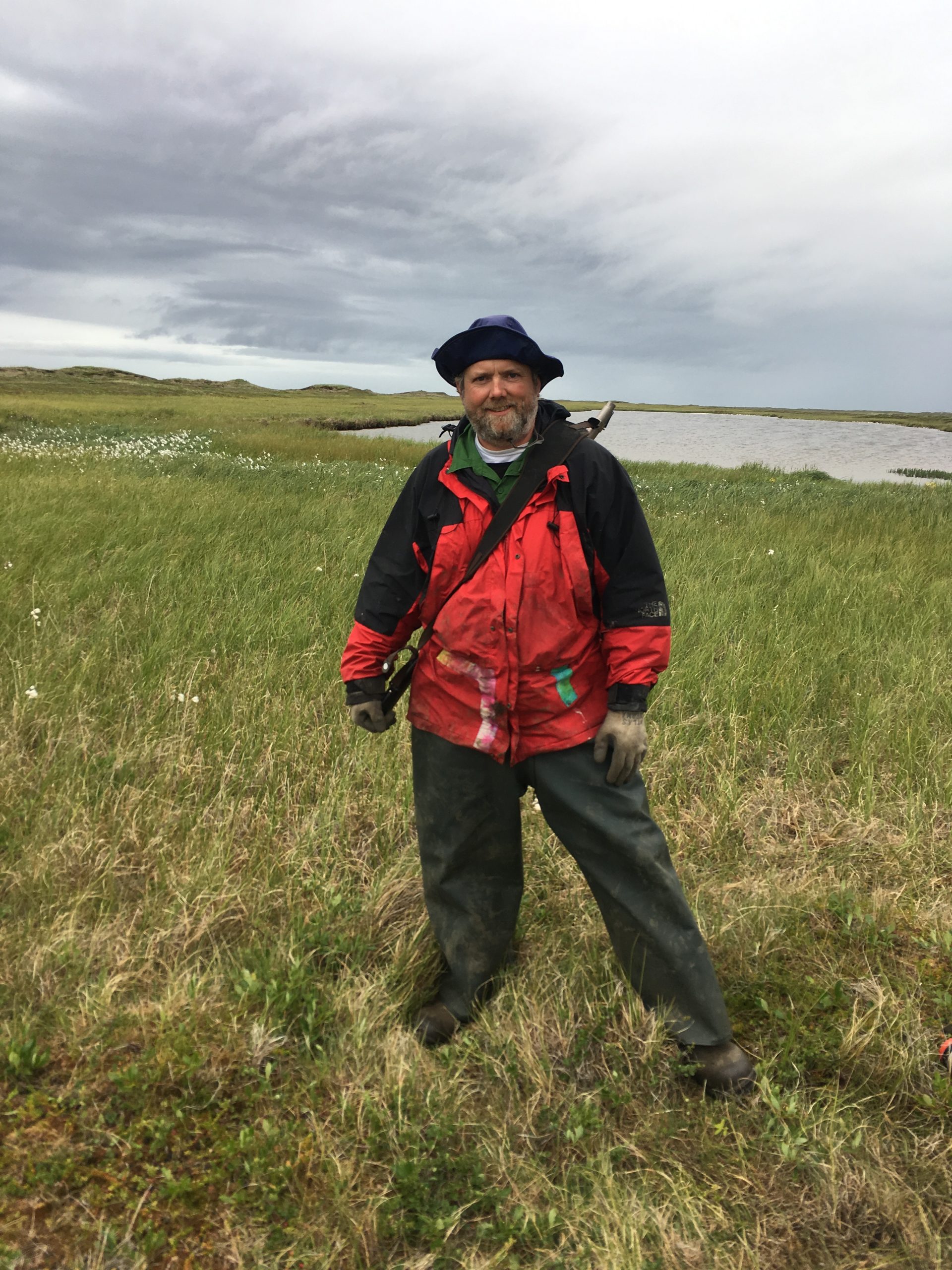 Chris stands in a grassy field with white flowers speckled througout the background, as well as a small pond in the background and a cloudy sky