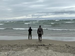 Two people stand centered in the photo, facing away from the camera. One is holding a Trimble GPS device and the other is walking towards them. They are on a beach with stormy-looking waters and grey skies.