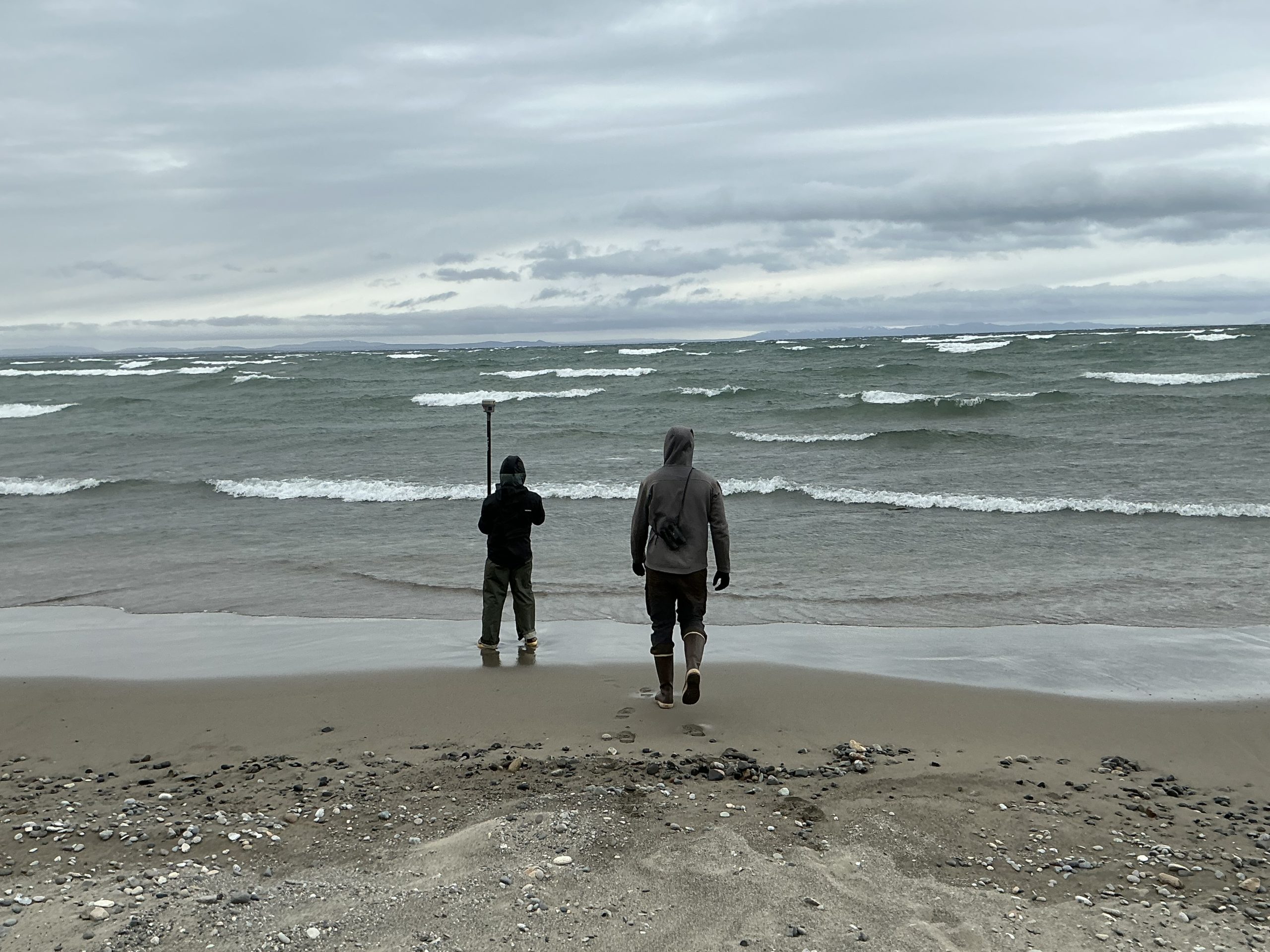 Two people stand centered in the photo, facing away from the camera. One is holding a Trimble GPS device and the other is walking towards them. They are on a beach with stormy-looking waters and grey skies.