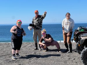 Four people standing on a beach smiling, one with thumbs up, one holding a peace sign, one croucing and doing a peace sign, and one is just smiling at the camera. The background shows a blue ocea and a clear blue sky.