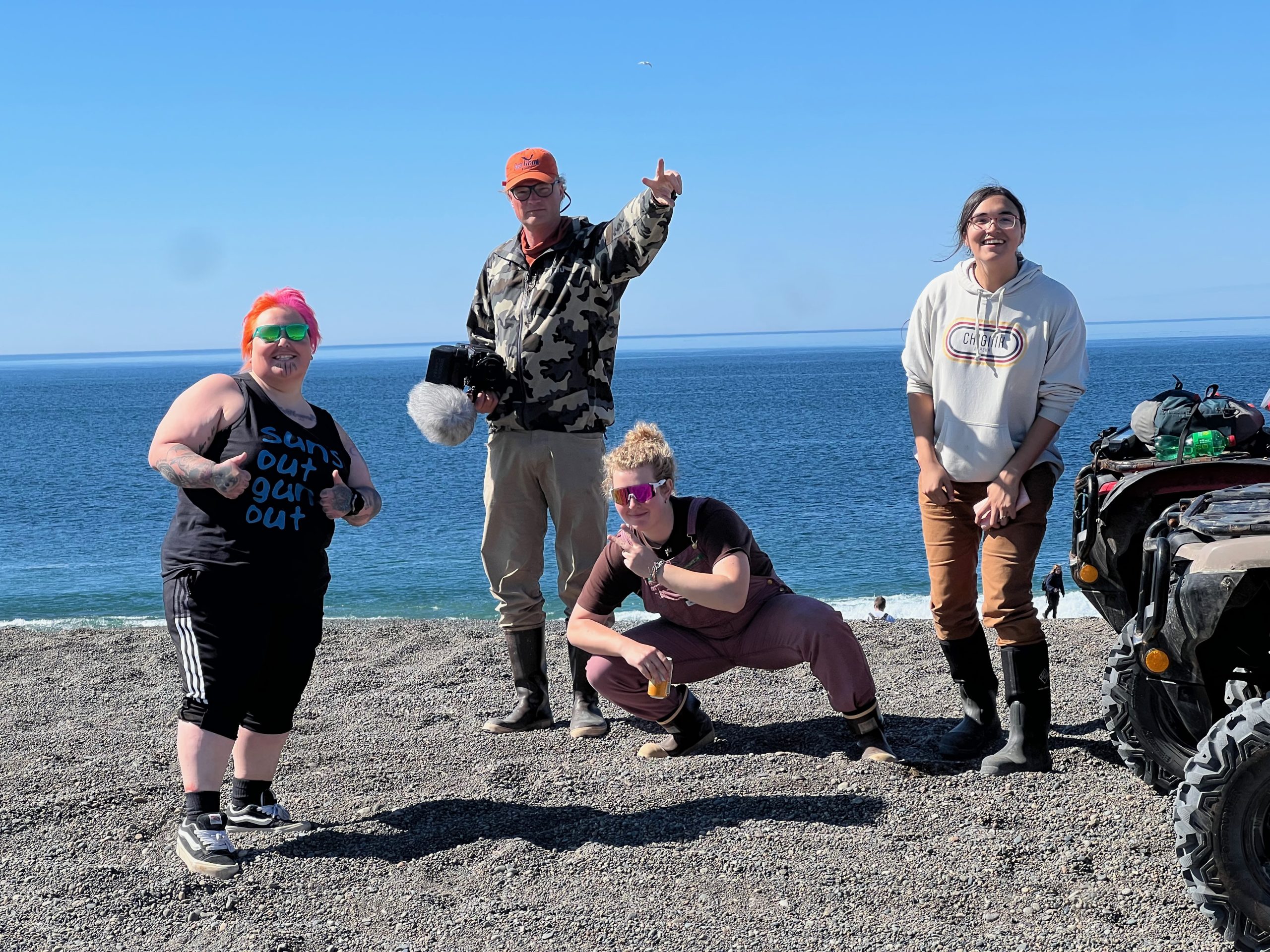 Four people standing on a beach smiling, one with thumbs up, one holding a peace sign, one croucing and doing a peace sign, and one is just smiling at the camera. The background shows a blue ocea and a clear blue sky.