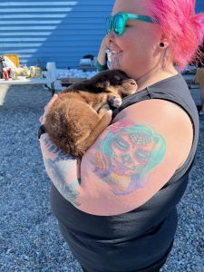 A puppy is snuggled against someone with bright pink hair and vibrant tattoos. In the background are some tables with food set up and a blue house.