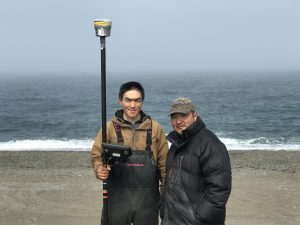 Two men from Gambell stand in the center, one is holding a Trimble instrument. The background shows a beach with blue water and a horizon that blends into a grey, misty sky.