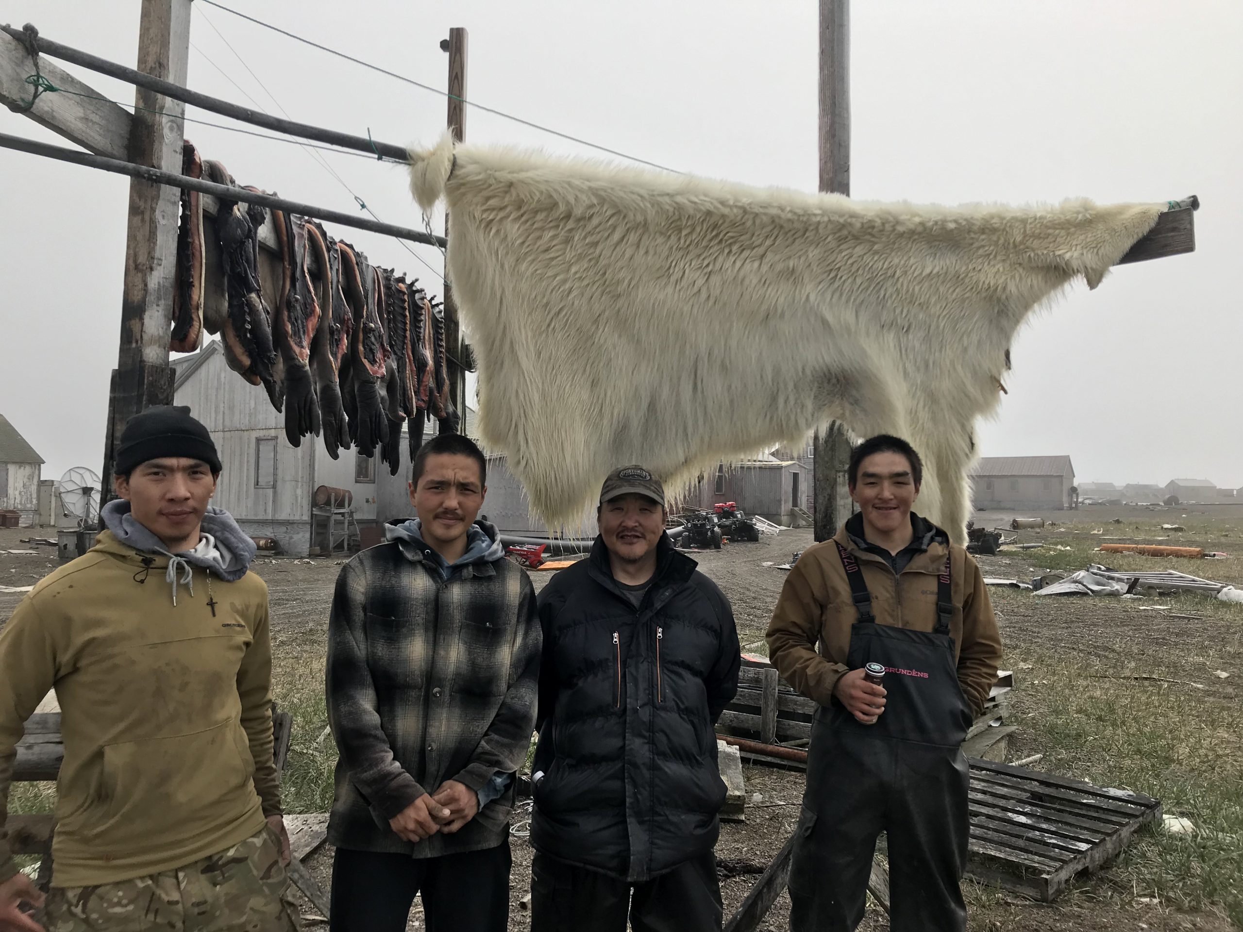 Four men from Gambell stand in front of a hanging polar bear skin and hanging seal skins. Behind them is a foggy Gambell village with a grey overcast day.