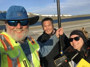 Three people stand close together for a selfie. One of them is holding a Trimble instrument. They are standing on a paved road. In the background there is a lake of dark blue water and a hill with patches of snow on it. The sky is blue and partially cloudy.
