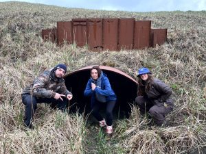 Three people squatting in front of a large storm drain tunnel in a grassy field. The grass is green and yellow, the sky is blue and a little cloudy.