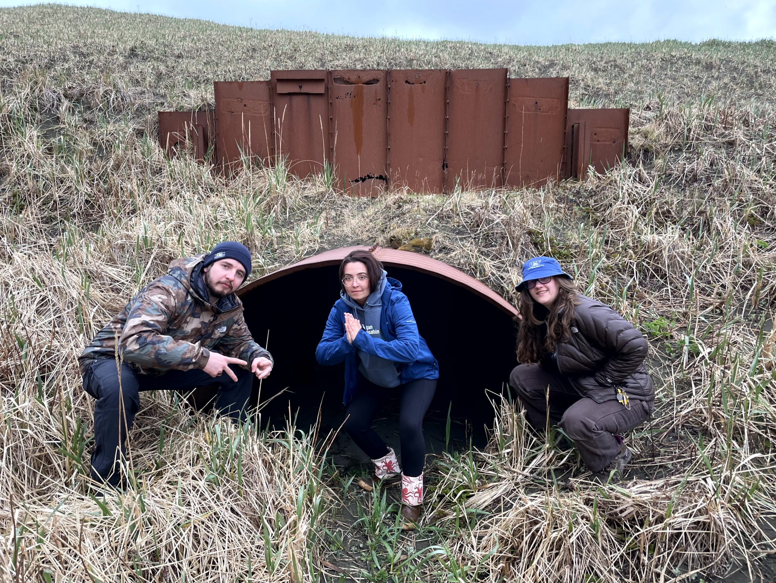 Three people squatting in front of a large storm drain tunnel in a grassy field. The grass is green and yellow, the sky is blue and a little cloudy.