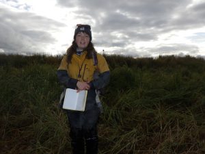 Someone stands facing the camera, holding a field notebook. They are standing in a field of tall grass, the sky is bright and cloudy
