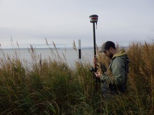 Someone stands on the right side of the image, their body is facing the left side of the image. They are standing in tall grass, holding a Trimble instrument. In the background there is calm ocean and an overcast sky.