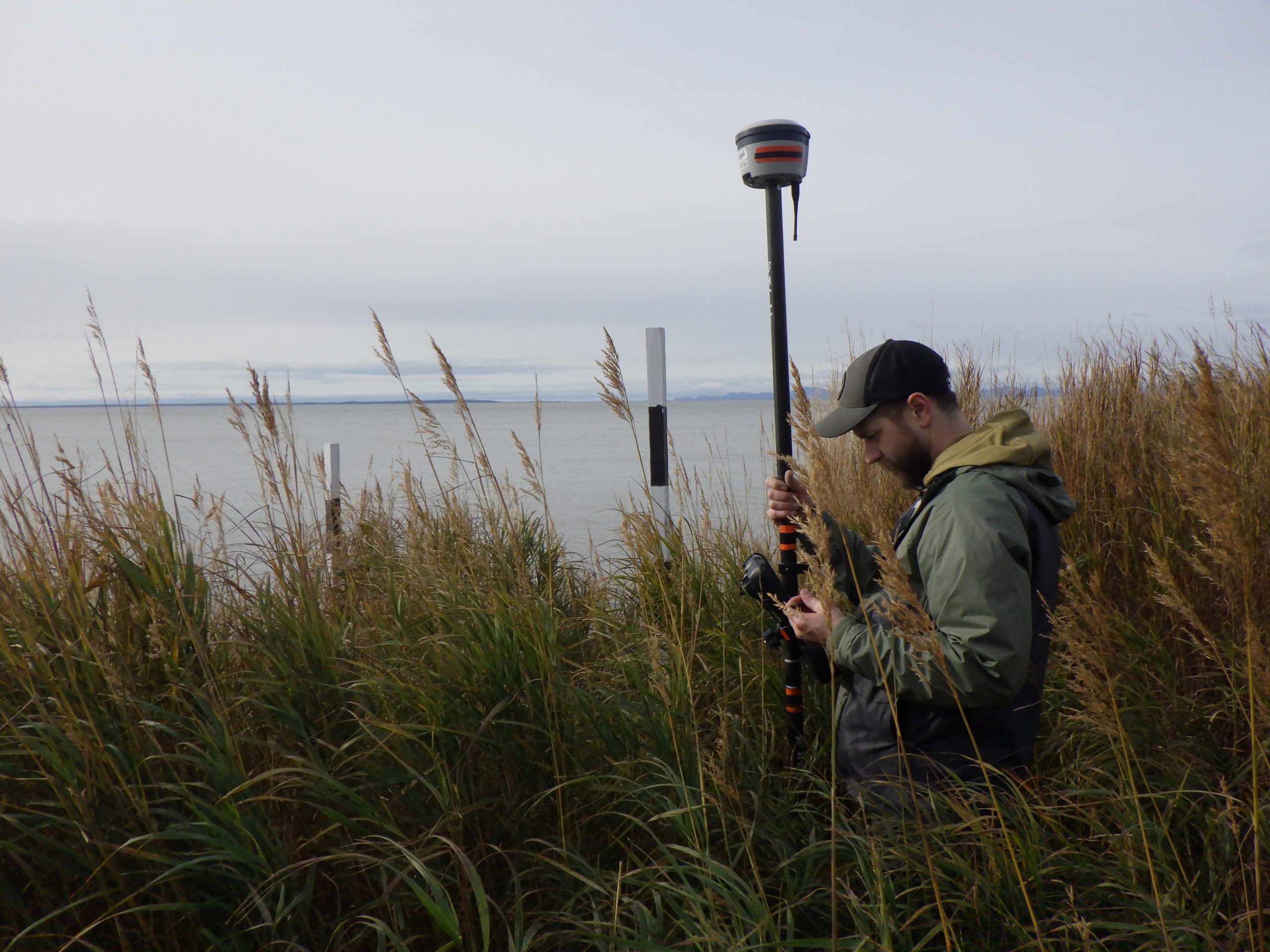 Someone stands on the right side of the image, their body is facing the left side of the image. They are standing in tall grass, holding a Trimble instrument. In the background there is calm ocean and an overcast sky.