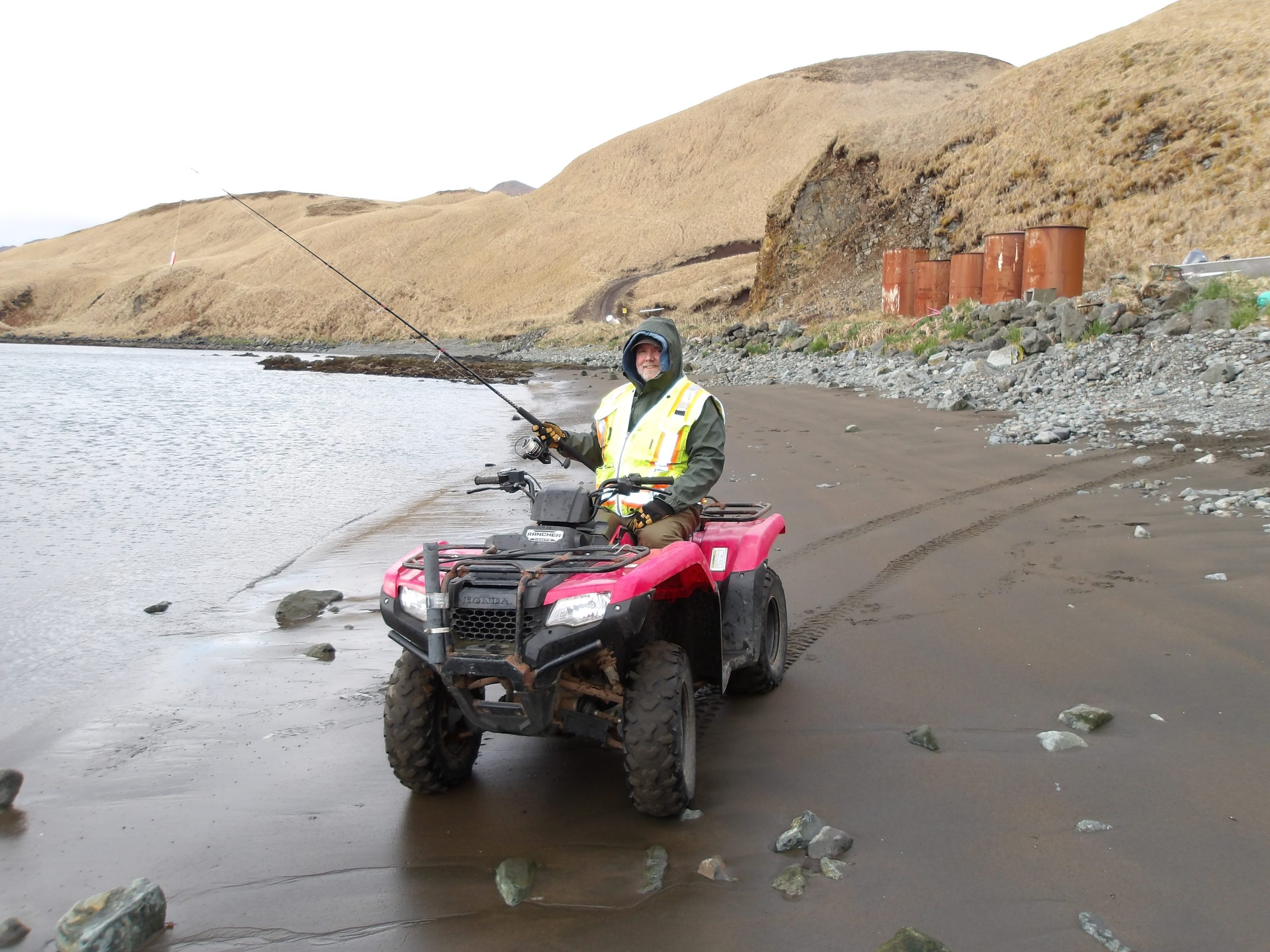 Chris sits on a red ATV on a beach, holding a fishing pole, the background is hilly with dry grass and an overcast sky
