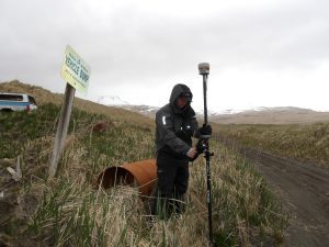 Someone stands near a rusted barrel, holding a Trimble instrument in a hilly grassy field. A sign next to them says "vehicle dump"