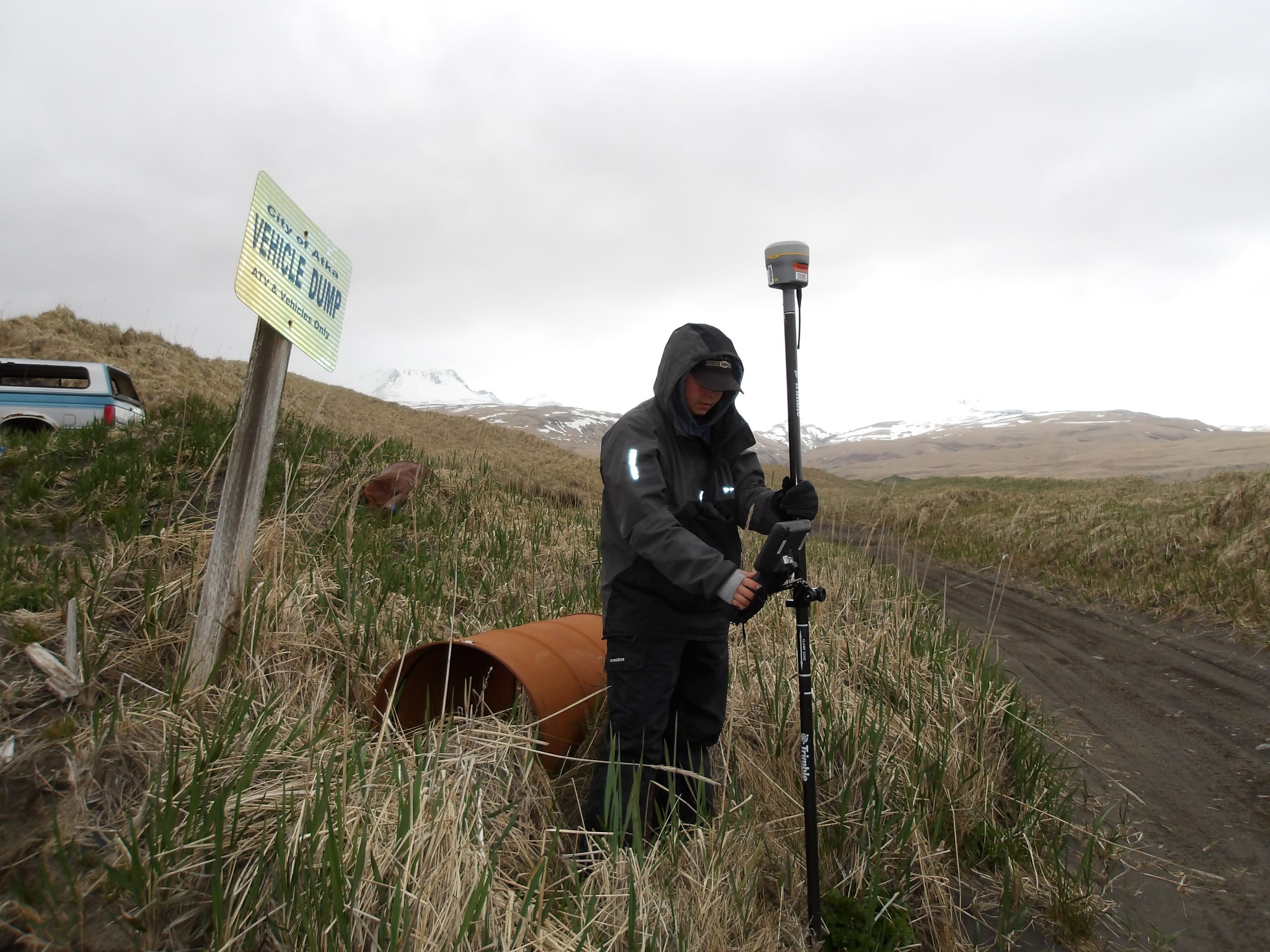 Someone stands near a rusted barrel, holding a Trimble instrument in a hilly grassy field. A sign next to them says "vehicle dump"