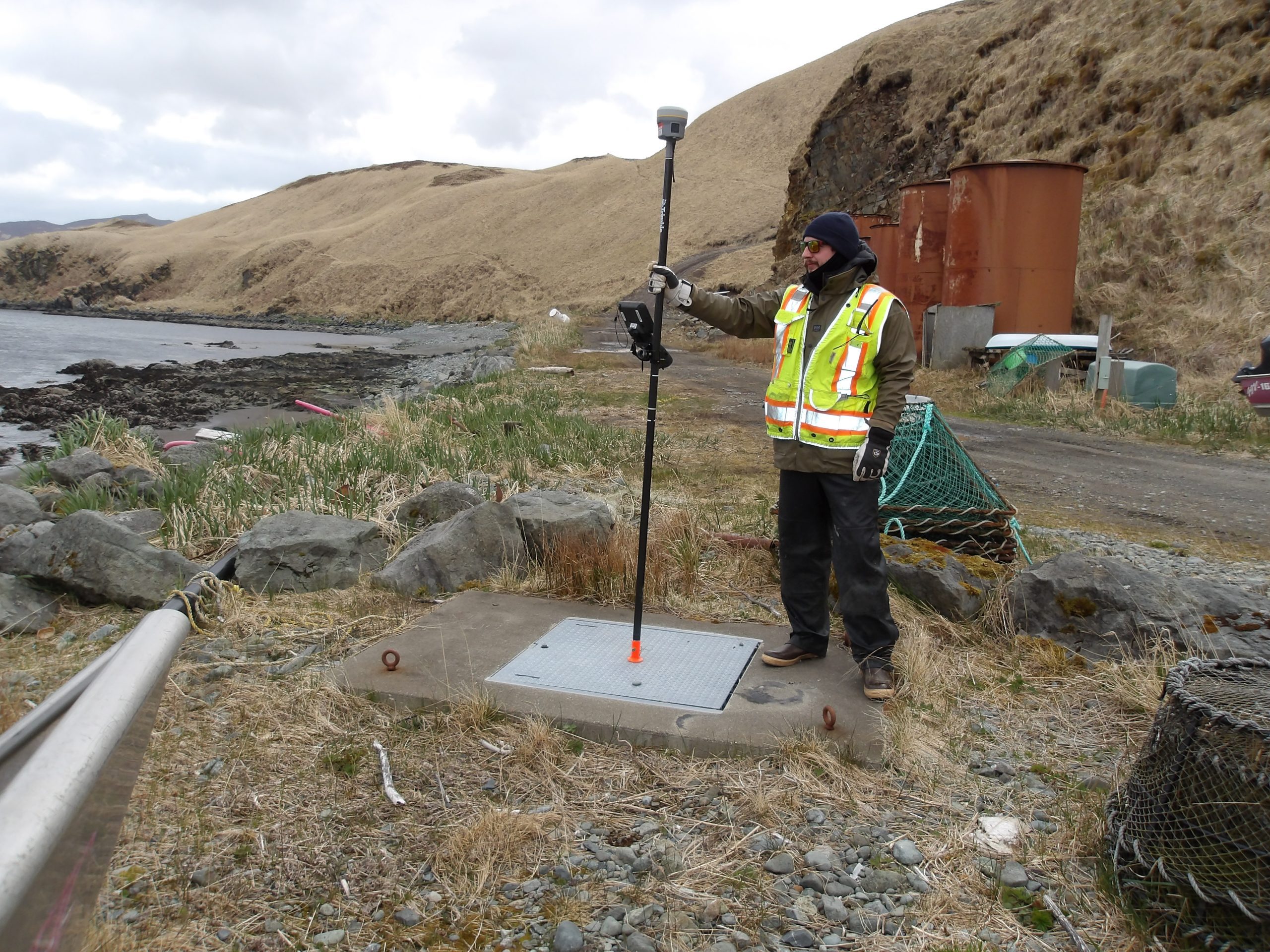 Someone stands with their Trimble instrument, on a hilly beach area. Some rusted cylinders are behind them.