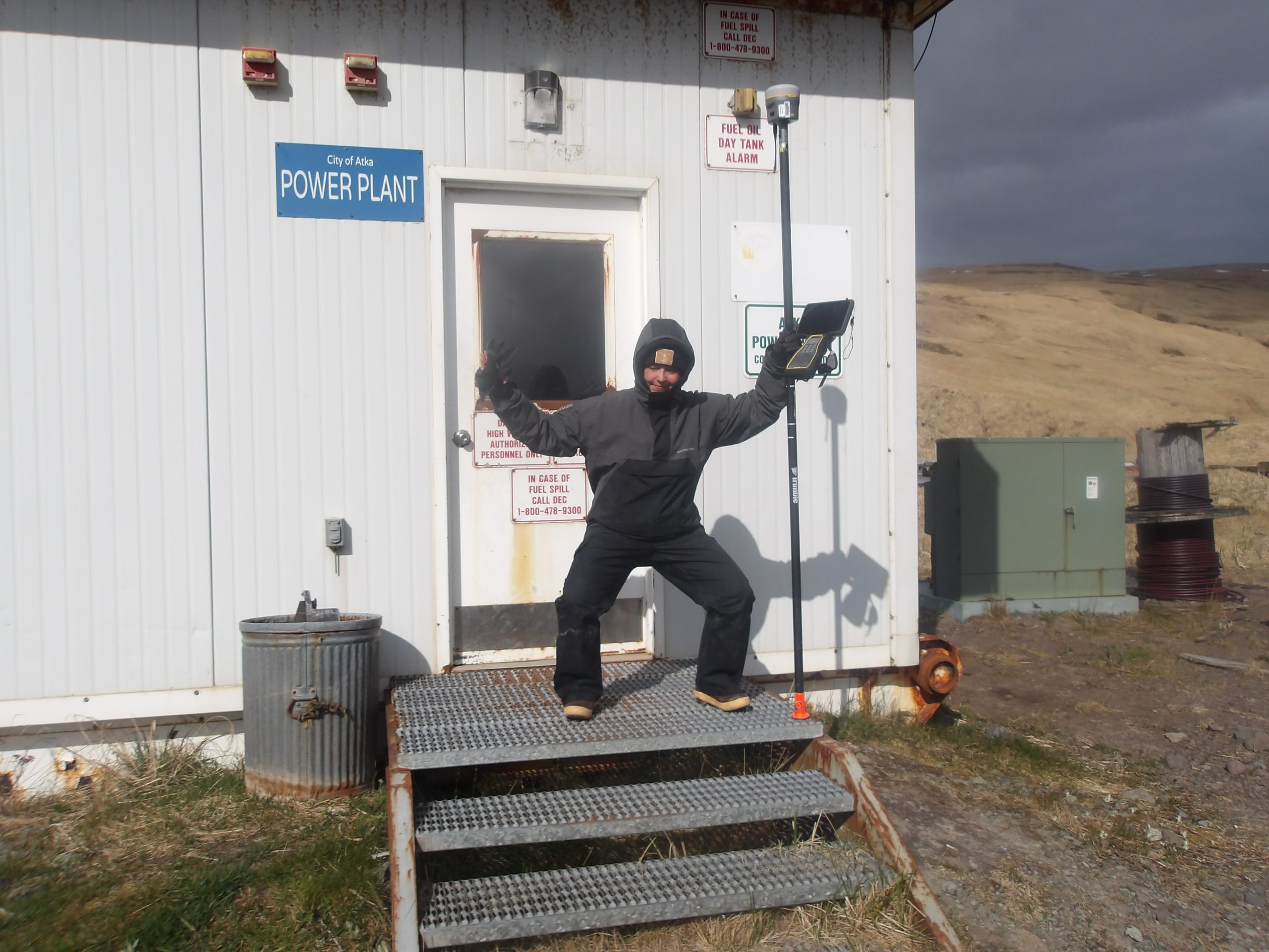 ACGL member stands in a half squat with their arms out, holding a Trimble instrument in front of a power plant building.