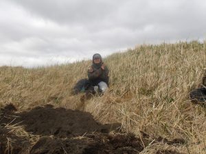 Someone sits on a small grassy hill, holding a sandwich and a peace sign, some fresh dirt is near their feet