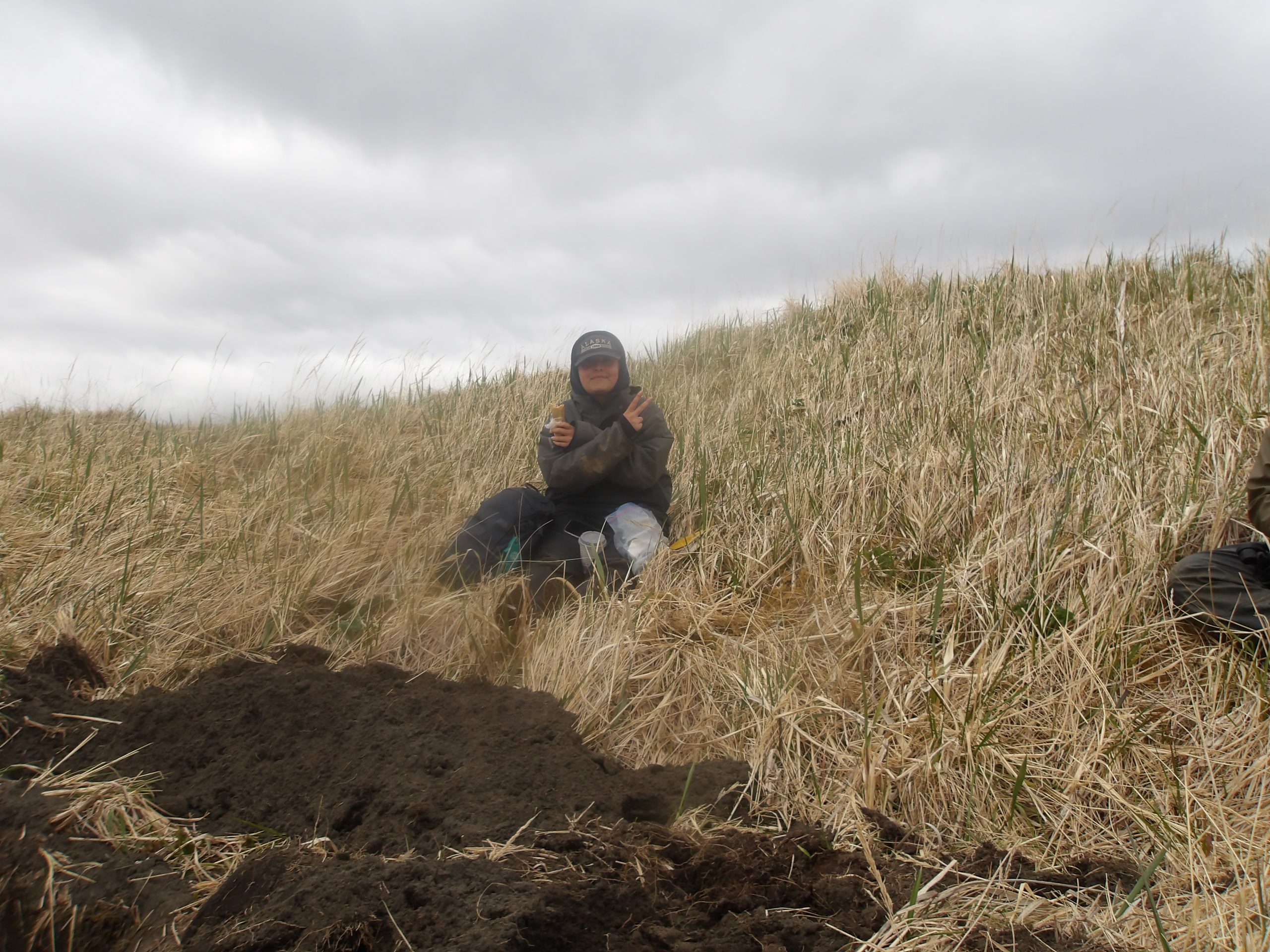 Someone sits on a small grassy hill, holding a sandwich and a peace sign, some fresh dirt is near their feet