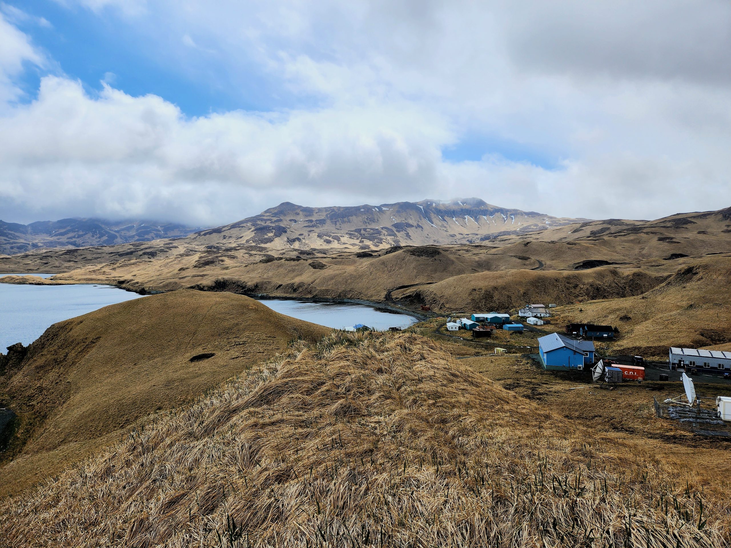 Hilly landscape with yellow grass, partially cloudy sky, and smooth waters. A little corner of Atka can be seen nestled in between the hills.