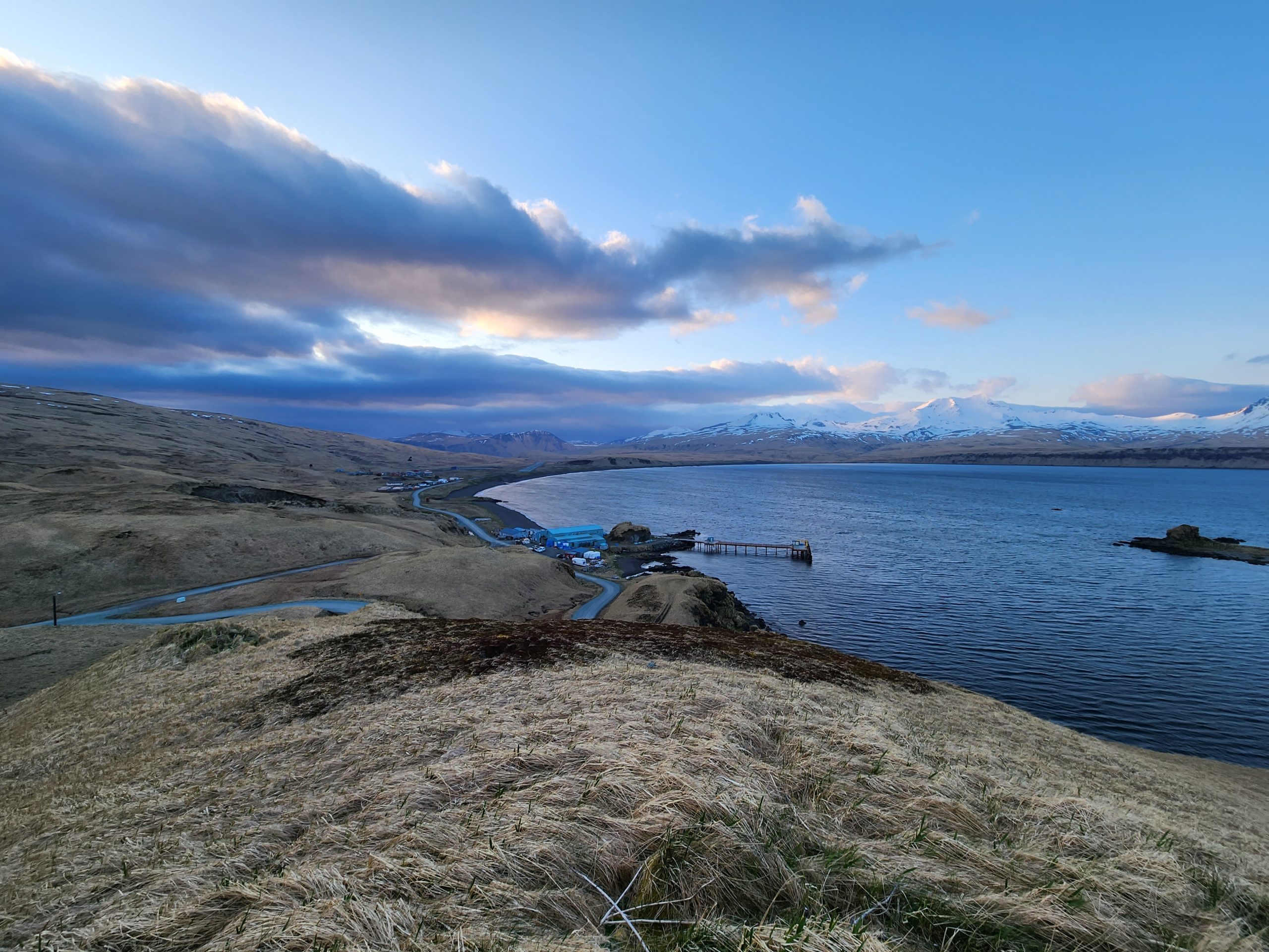 Another landscape with rolling grassy hills, showing a different angle of Atka with smooth blue waters, and a partially cloudy sky with blues and pinks. A snow-capped mountain pokes out of the background.