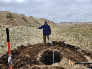 Chris stands behind a freshly dug hole, behind him is grassy hills and an overcast sky.