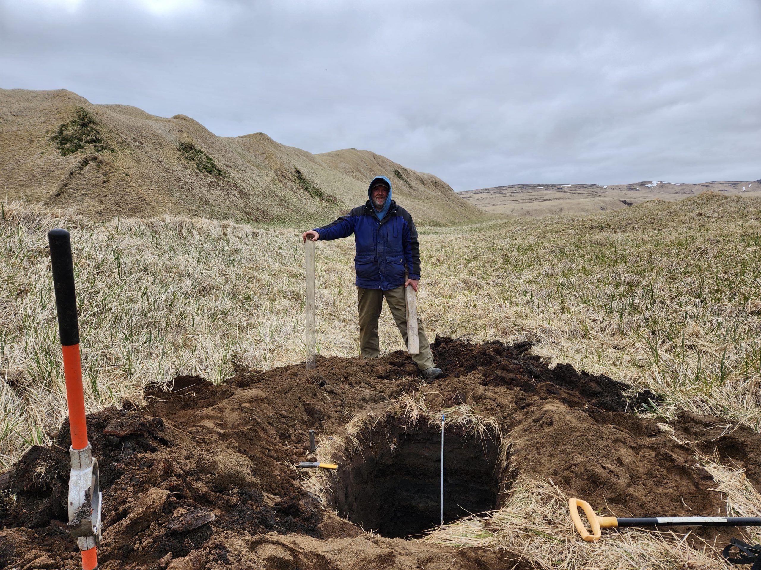 Chris stands behind a freshly dug hole, behind him is grassy hills and an overcast sky.
