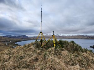 Two Trimble homing instruments stand on a grassy hill, the background shows a calm body of water, snow capped mountains and low, overcast clouds.