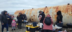 A group of people stand around tables coverd in an assortment of food. They are standing on gravelly ground. Behind them is a large, partially rusted cylinder. The sky is overcast.