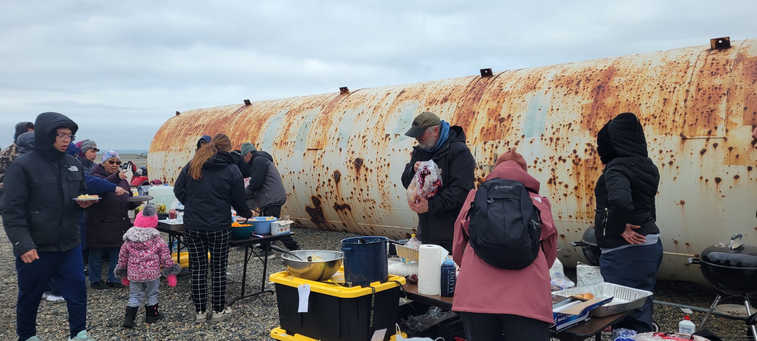 A group of people stand around tables coverd in an assortment of food. They are standing on gravelly ground. Behind them is a large, partially rusted cylinder. The sky is overcast.