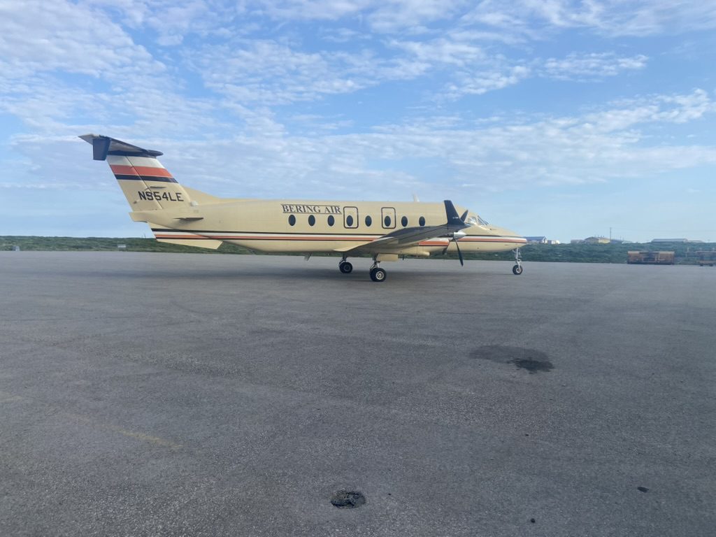 An airplane sits on a runway. The plane says "Bering Air". There is some brush and houses along the horizon. The sky is blue and speckled with clouds.