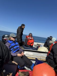 A group of people sitting in boats, some are wearing life jackets. They are sat by the back of the boats. The water is blue and calm. The sky is blue and nearly clear of clouds.