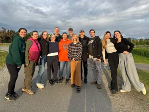 People standing near each other for a group photo. They are standing on a paved road. The area is grassy and looks like a park. The sky is cloudy and overcast.
