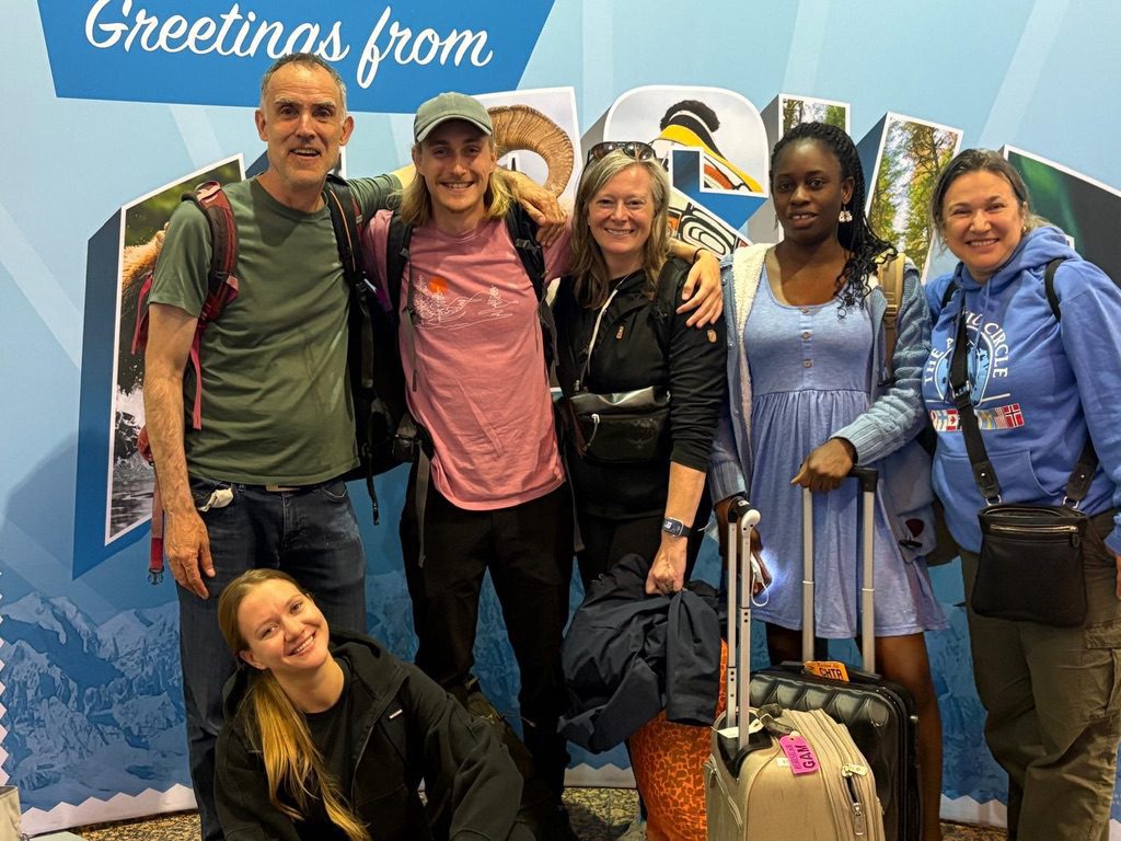 People standing near each other for a group photo. Behind them is a sign that says "Greetings from Alaska".