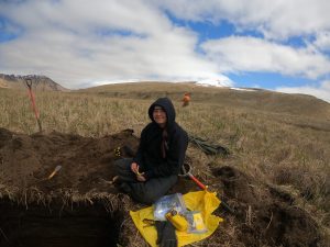 Someone sits next to a freshly dug hole, the background shows grassy rolling hills and a partially cloudy sky.