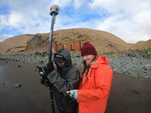 Two people stand on a beach with a Trimble instrument, behind them are dried-grassy hills