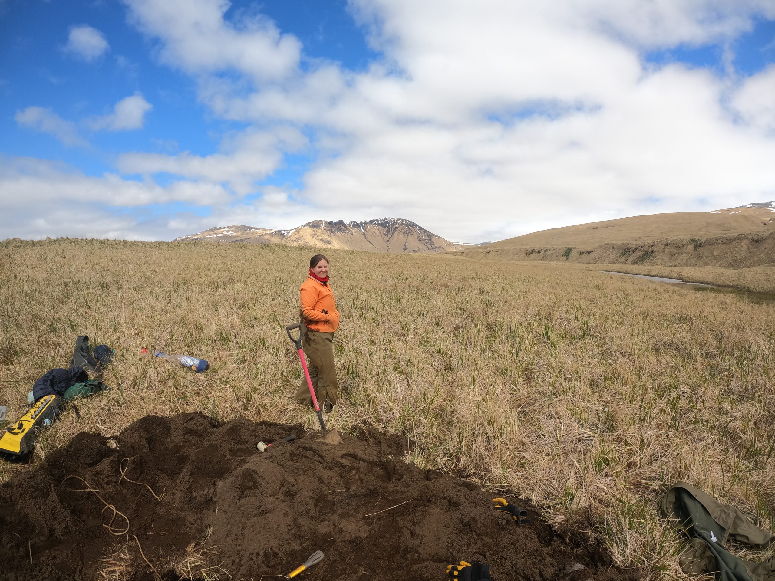Someone stands next to fresh dirt in a yellow-grassy field. The sky is partially cloudy.