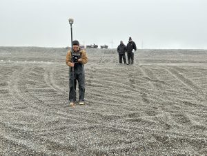 Someone stands on a gravelly beach with tire tracks, holding a Trimble instrument. Behind them are two other people looking towards the person holding the Trimble. There are a couple of ATV's sitting on the horizon, as well as a couple of windmills visible in the haze. The sky is grayish-white.