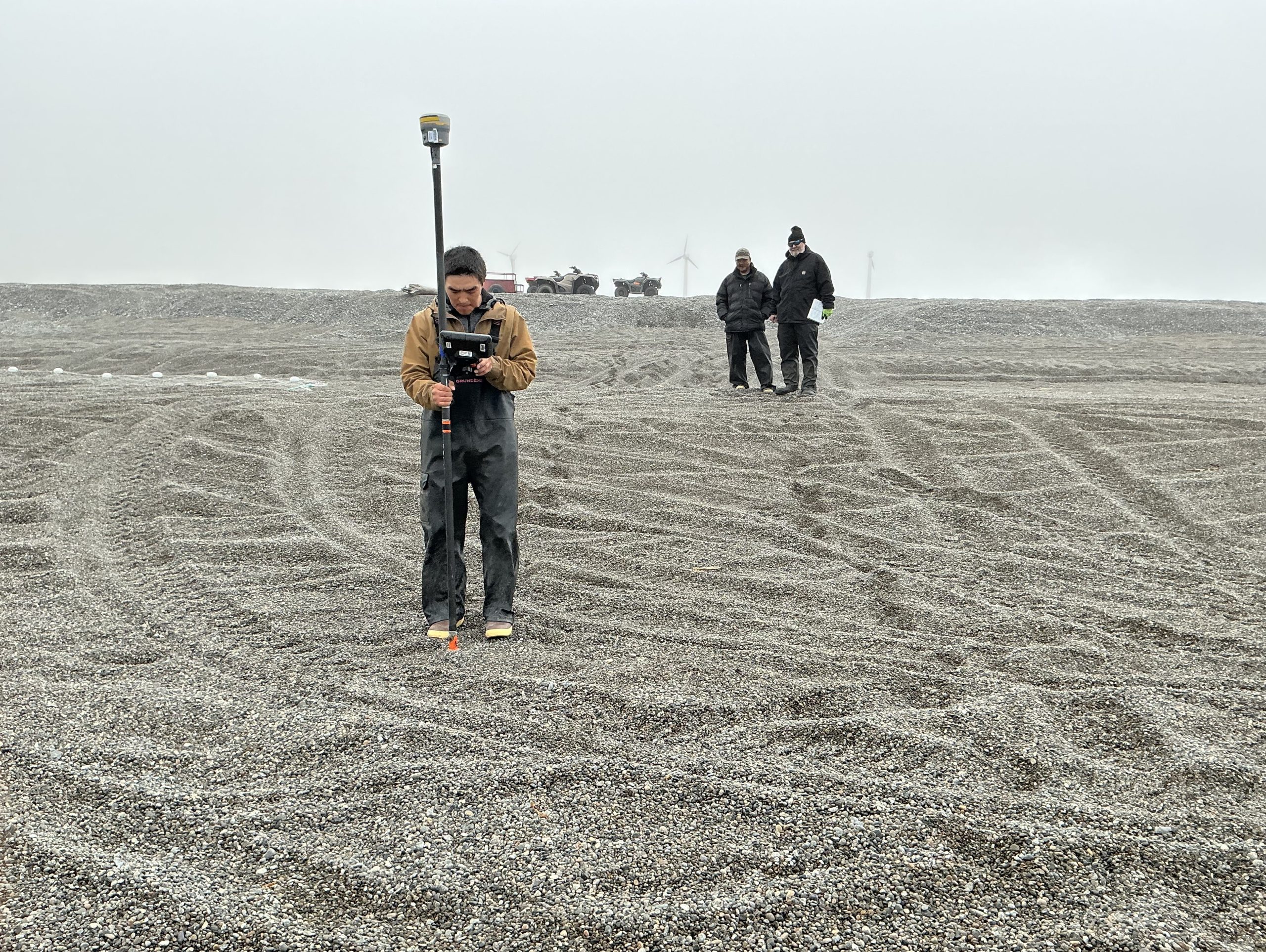 Someone stands on a gravelly beach with tire tracks, holding a Trimble instrument. Behind them are two other people looking towards the person holding the Trimble. There are a couple of ATV's sitting on the horizon, as well as a couple of windmills visible in the haze. The sky is grayish-white.