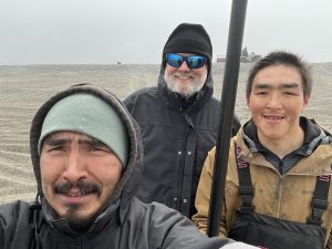 Three people stand together for a selfie. One of them is holding a metal pole. They are in a grey gravelly field covered in tire tracks. There is someone standing next to an ATV and a trailer on the horizon. The sky is grey.