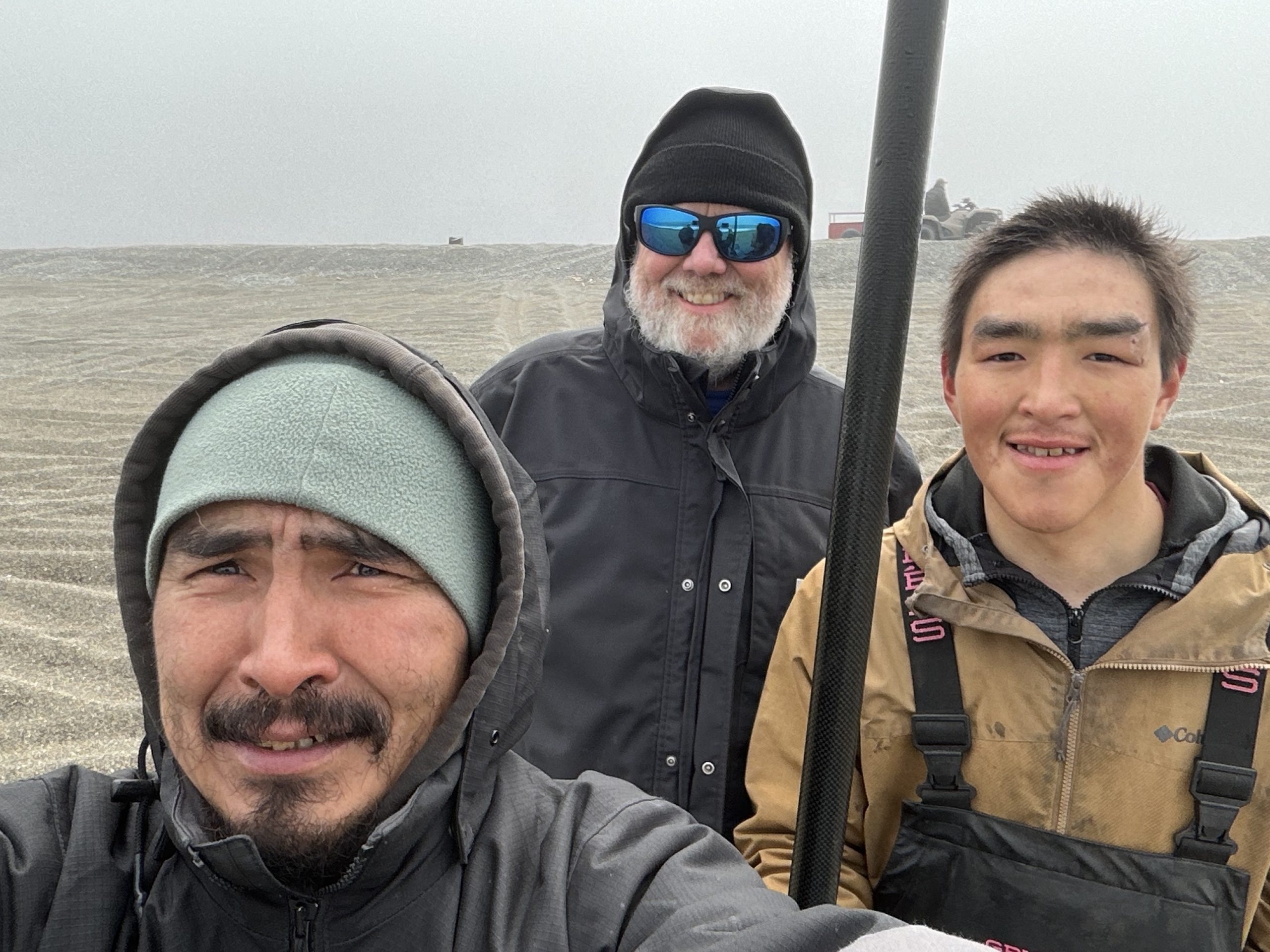 Three people stand together for a selfie. One of them is holding a metal pole. They are in a grey gravelly field covered in tire tracks. There is someone standing next to an ATV and a trailer on the horizon. The sky is grey.